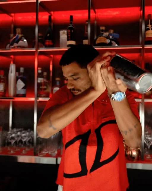 Bartender do BRABO a agitar uma coqueteleira em frente ao bar iluminado a vermelho, cheio de garrafas